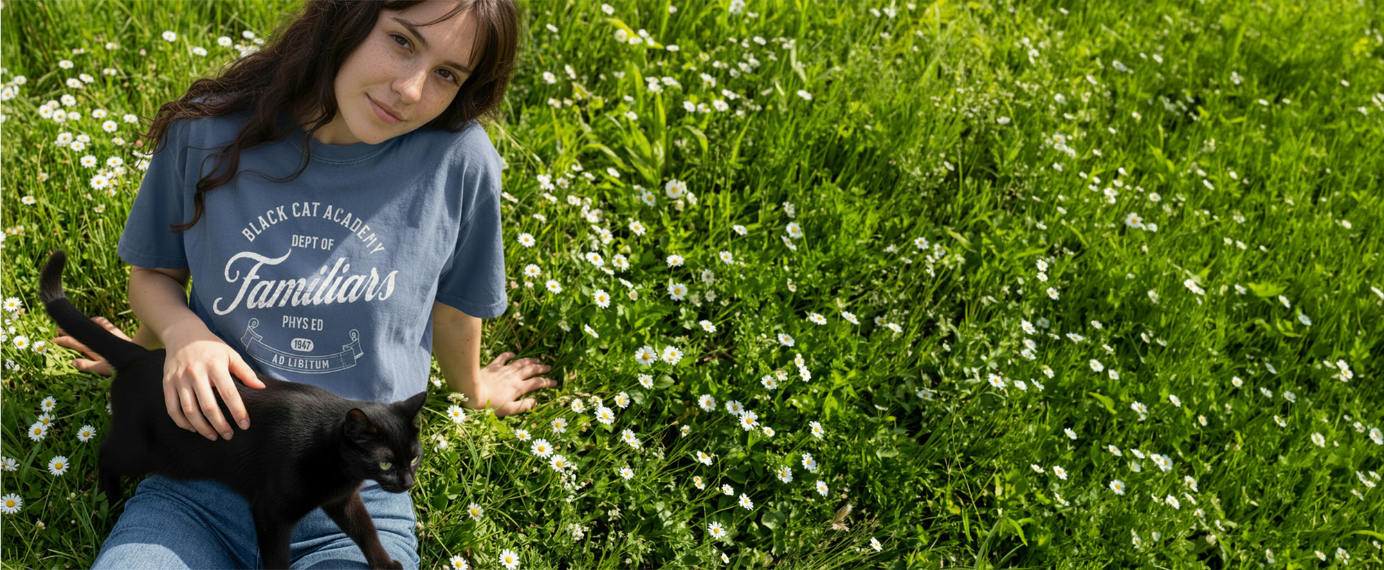 Woman wearing a Black Cat Academy tee, holding a black cat in a grassy field with white flowers